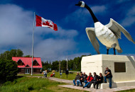 group of people sitting at the base of a large white goose monument with Canada flag flying nearby